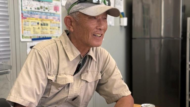 工場の休憩室で書類を手に笑顔で話す作業服の株式会社中友農産 中村 友平さん