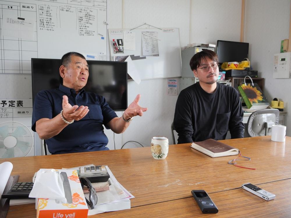 会議室で2人の男性が机を挟み、左の男性が身振りで説明している写真