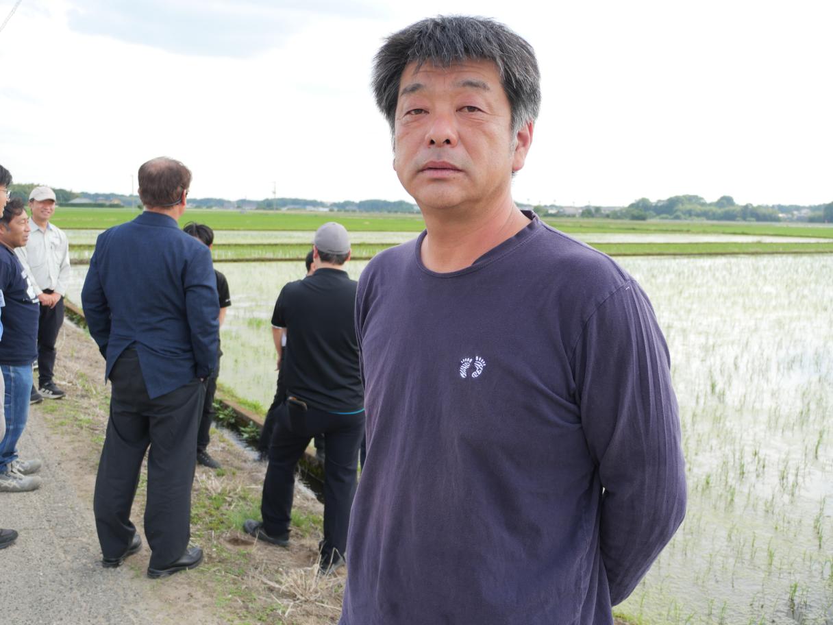 田んぼの脇で立つ男性、後ろに数人が集まる写真