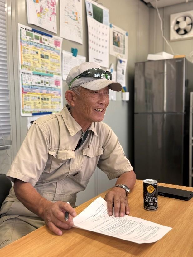 工場の休憩室で書類を手に笑顔で話す作業服の株式会社中友農産 中村 友平さん