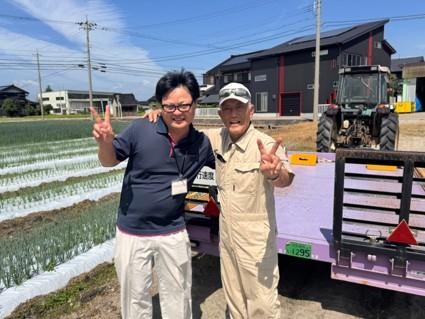 田んぼ脇でトラック前に立ち、笑顔でピースする株式会社中友農産 中村 友平さん